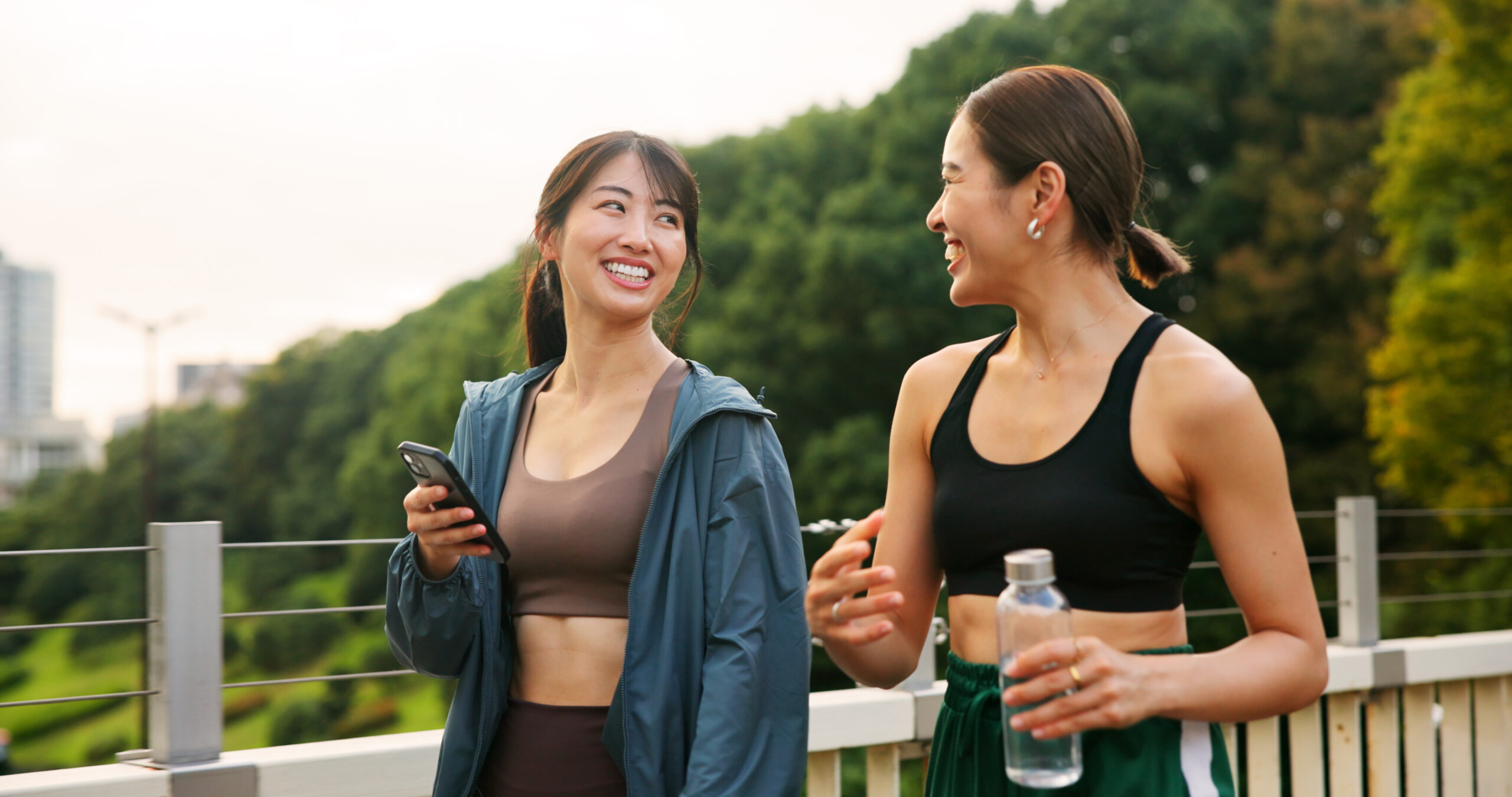 Fitness, happy friends and girls walking at park, talking or communication on phone. Japanese women, sports and outdoor together for body health conversation, exercise or gossip on break with bottle.